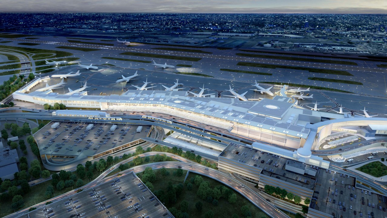 Aerial view of a modern airport terminal with multiple airplane gates, a parking lot, and surrounding roads, illuminated at dusk.