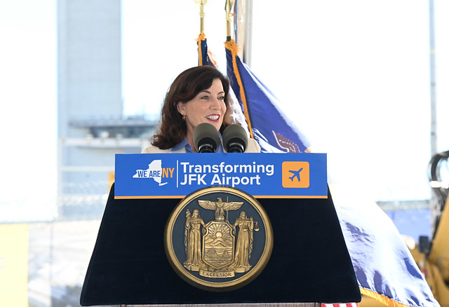 A woman speaks at a podium promoting the transformation of JFK Airport, featuring the state seal and a logo.